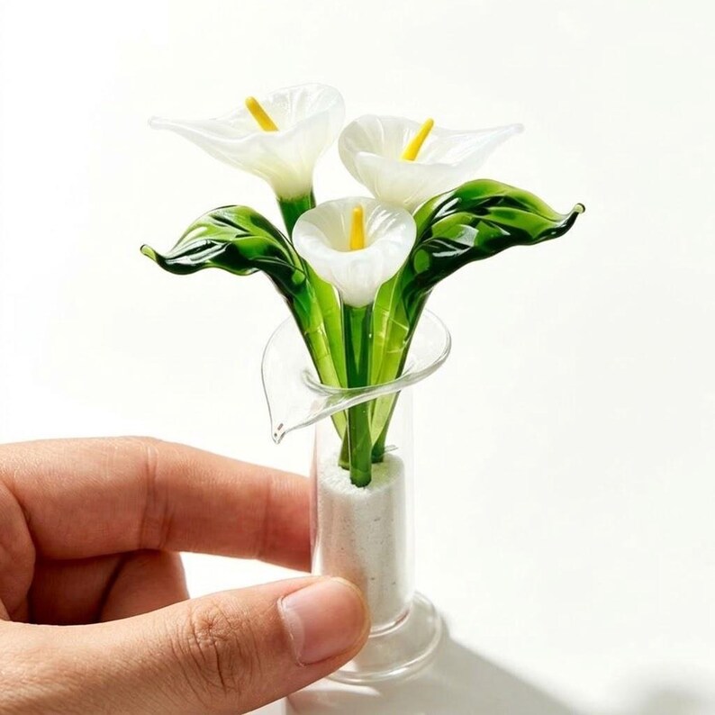 May include: A glass sculpture of three white calla lilies with yellow centers and green stems and leaves, arranged in a clear glass vase filled with white sand. The sculpture features smooth, translucent surfaces and delicate, curved petals.