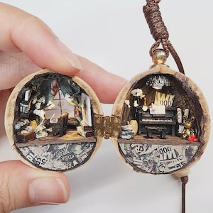 May include: A miniature diorama inside a hinged walnut shell pendant on a brown cord. The left side features a room with a window, curtains, and furniture. The right side contains a black piano, a small golden rabbit figure, and a light fixture.
