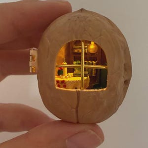以下が含まれることがあります： 中をくり抜いたクルミの殻の中に収められたミニチュアのジオラマ。テーブル、本、温かみのある室内照明を備えた小さな部屋が特徴です。殻には、十字の桟が入った金色の金属製窓枠と、側面に小さな金属製の蝶番が付いています。