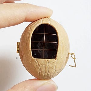May include: A hollowed-out walnut shell featuring a small, rectangular opening covered by a thin, dark brown wire grid. The shell is equipped with a small gold-toned metal hinge on the left side and a tiny metal hook closure on the right side.