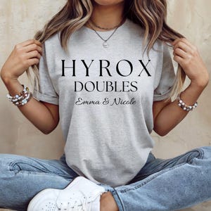 May include: A light gray t-shirt with the words "HYROX DOUBLES" in a bold, black font. Below, in a smaller font, are the names "Emma & Nicole". The shirt is a casual style.