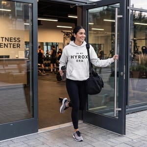 May include: A light gray hooded sweatshirt with the black text IN MY HYROX ERA printed on the front, paired with black leggings, gray athletic sneakers, and a black duffel bag. The garment features a front kangaroo pocket and long sleeves.