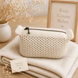 May include: A cream-colored, rectangular crochet pouch with a textured, honeycomb-like stitch pattern. The pouch features a dark metal zipper closure with a small spherical pull and two small, protruding side tabs at the top corners.