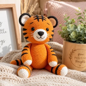 Può includere: Un giocattolo a forma di tigre all'uncinetto arancione e nero, con dettagli bianchi. La tigre ha strisce nere e un'espressione sorridente. Sullo sfondo, una stampa incorniciata con la scritta "Enjoy the little things".