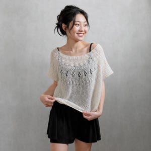 Peut inclure: Un haut en tricot couleur crème à manches courtes, doté d'un motif en dentelle ajourée complexe sur la poitrine et les épaules, d'une large encolure arrondie et de bords côtelés au col et aux manches, porté sur un caraco et un short noirs.