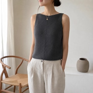 May include: A dark charcoal gray sleeveless knit top featuring a boat neckline and a subtle vertical seam detail running down the center front. The garment is paired with light beige high-waisted trousers that have a button closure and pleats.