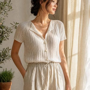 May include: A short-sleeved, off-white knit cardigan featuring a ribbed texture, a V-neckline, and three small, round, pearlescent buttons down the front. The garment is paired with light beige, high-waisted trousers with a gathered elastic waistband.