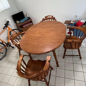 May include: A wooden dining set featuring an oval table and four chairs. The table and chairs are made of light brown wood. Two chairs have armrests. The set is placed on a tiled floor.