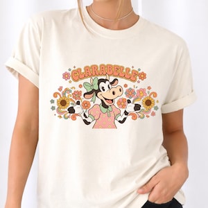 May include: Cream-colored t-shirt featuring a cartoon cow character named Clarabelle. The cow is wearing a pink dress and a green bow, surrounded by colorful flowers and the name "CLARABELLE" in orange letters. The shirt has rolled-up sleeves.