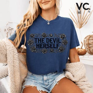 Puede incluir: Una camiseta azul marino con el texto "THE DEVIL HERSELF" en una fuente gótica, enmarcada por un diseño de vid espinosa y rosas. La camiseta es de corte holgado y tiene mangas cortas. La imagen también muestra a una persona que lleva la camiseta.