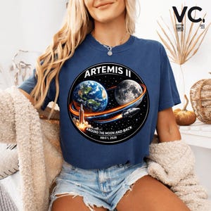 May include: Navy blue t-shirt featuring a circular graphic with the text "ARTEMIS II" and "AROUND THE MOON AND BACK April 1, 2026." The graphic depicts Earth, the Moon, a rocket, and a spacecraft. The shirt also has the text "VC CUSTOM EDITIONS SINCE 2023."