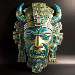 Puede incluir: Una máscara azteca detallada, en tonos turquesa, dorado y azul, con una expresión feroz. La máscara presenta intrincadas tallas, cuernos y adornos para las orejas. Es un objeto decorativo, probablemente para colgar en la pared.