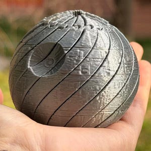 May include: A silver-colored, spherical object with detailed surface patterns and a circular indentation. The object has a textured appearance with diagonal lines and small rectangular details, resembling a Death Star model.