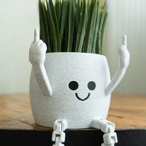 Puede incluir: Una maceta de cerámica blanca moteada con una cara de dibujos animados, brazos y piernas. La maceta tiene una boca sonriente y está haciendo la señal del dedo medio con ambas manos. Está llena de hierba artificial verde.