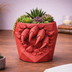 May include: A red planter with a claw design, filled with various succulents. The planter has a geometric shape and a textured surface. The claw design appears to be breaking through the planter's surface.