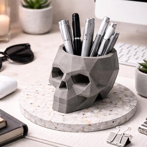 Puede incluir: Un portalápices gris en forma de calavera de hormigón lleno de bolígrafos plateados y negros. El portalápices se encuentra sobre un posavasos redondo y moteado. Otros artículos en el escritorio incluyen gafas de sol, un reloj y una pequeña suculenta en una maceta gris.