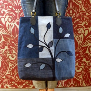 May include: A blue denim tote bag with a patchwork design featuring a stylized tree with leaves. The bag has black leather straps and a top zipper closure.