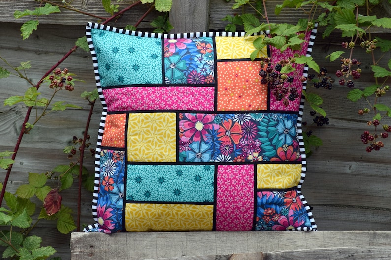 May include: A colorful patchwork pillow with a black and white trim. The pillow is made of different fabrics with floral patterns in shades of pink, yellow, blue, and green.