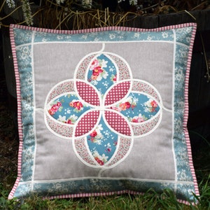 K&ouml;nnte beinhalten: Ein quadratisches Kissen mit einem Blumendesign in den Farben Rosa, Blau und Wei&szlig;. Das Kissen hat einen hellgrauen Hintergrund und einen blau-wei&szlig; gestreiften Rand.