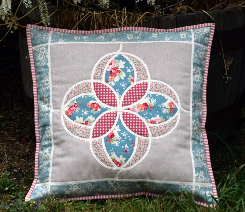 K&ouml;nnte beinhalten: Ein quadratisches Kissen mit einem Blumendesign in den Farben Rosa, Blau und Wei&szlig;. Das Kissen hat einen hellgrauen Hintergrund und einen blau-wei&szlig; gestreiften Rand.
