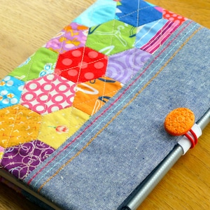 Könnte beinhalten: Ein farbenfrohes Patchwork-Stoff-Notizbuch mit einem Denim-Umschlag und einem Stifthalter. Das Notizbuch hat einen leuchtend orangefarbenen Knopfverschluss.
