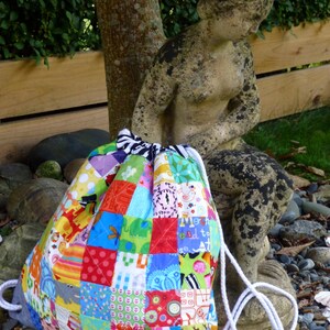 May include: A colourful patchwork drawstring bag with a white cord. The bag is made of various fabrics with different patterns and colours, including red, blue, green, yellow, and pink. The bag is sitting on a bed of rocks and shells.