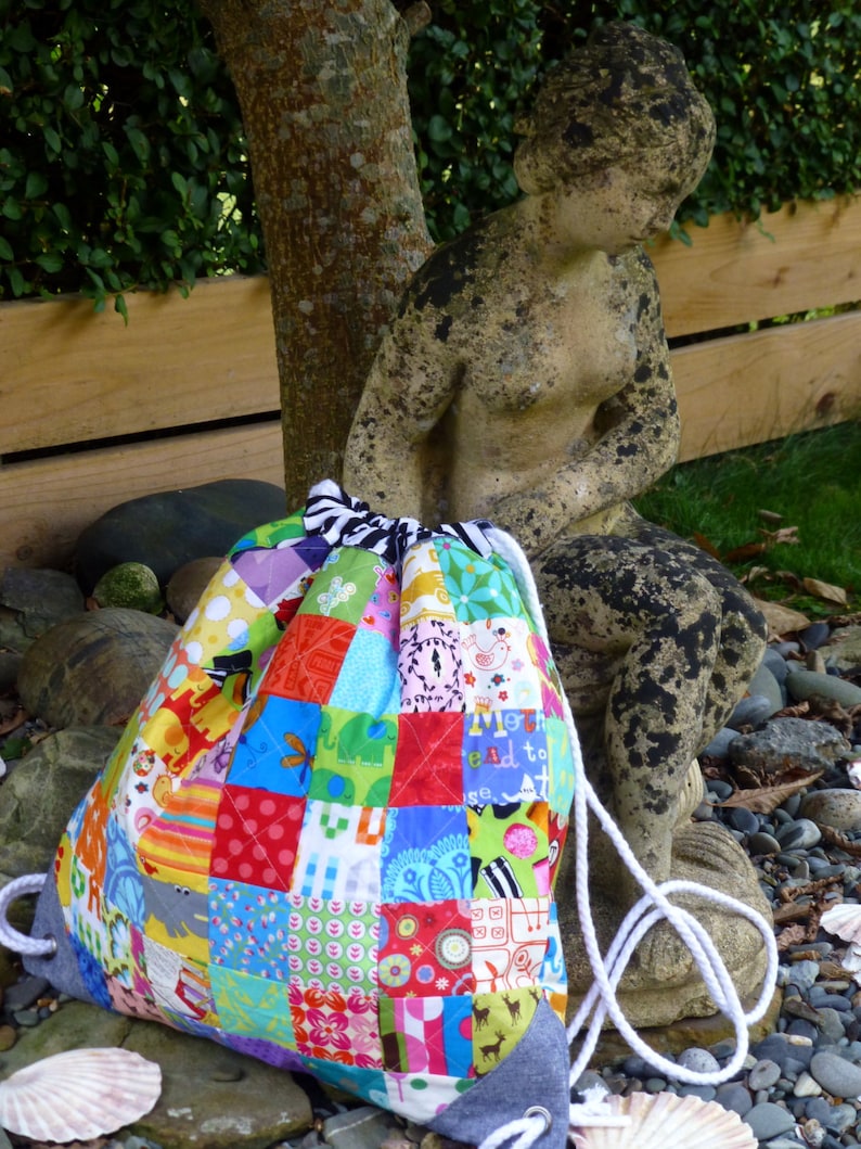 May include: A colourful patchwork drawstring bag with a white cord. The bag is made of various fabrics with different patterns and colours, including red, blue, green, yellow, and pink. The bag is sitting on a bed of rocks and shells.