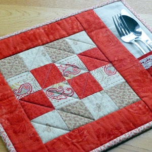 May include: A red and cream quilted placemat with a pocket for silverware. The placemat has a patchwork design with paisley and floral patterns.