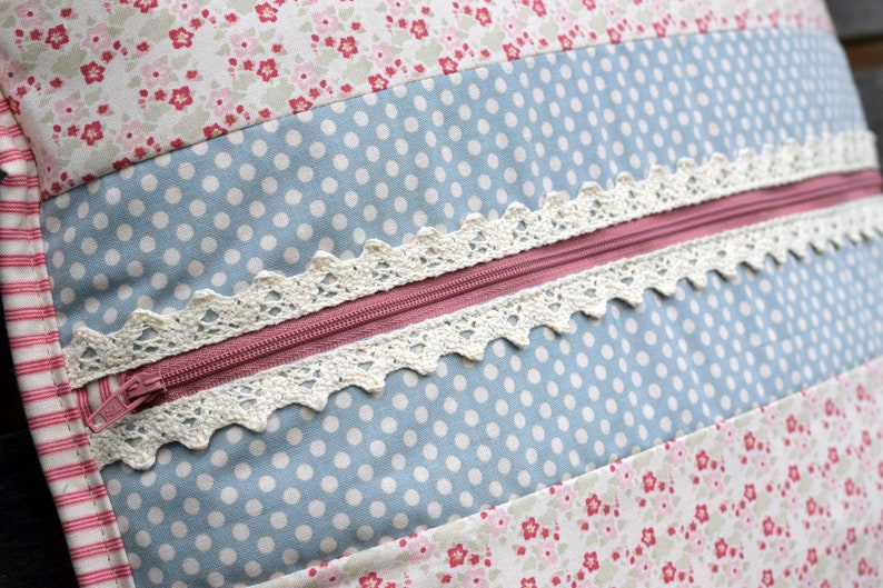 K&ouml;nnte beinhalten: Nahaufnahme eines Kissens mit Rei&szlig;verschluss und Spitzenbesatz. Das Kissen besteht aus Patchwork-Stoff mit einem blau-wei&szlig;en Punkte-Muster, einem rosa-wei&szlig;en Blumenmuster und einem wei&szlig;-rosa gestreiften Muster.