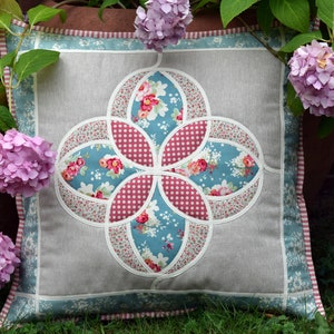 K&ouml;nnte beinhalten: Ein dekoratives Kissen mit einem floralen Muster in Rosa-, Blau- und Wei&szlig;t&ouml;nen. Das Kissen ist von rosa Hortensien umgeben.