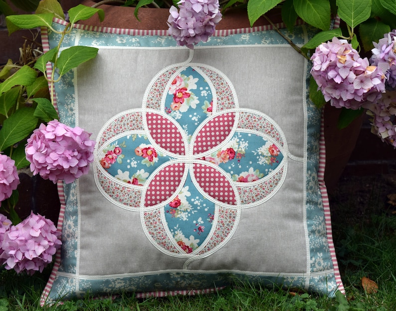 K&ouml;nnte beinhalten: Ein dekoratives Kissen mit einem floralen Muster in Rosa-, Blau- und Wei&szlig;t&ouml;nen. Das Kissen ist von rosa Hortensien umgeben.
