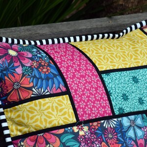 May include: A colorful patchwork pillow with a black and white striped border. The pillow is made of different fabrics with floral and geometric patterns in shades of pink, yellow, blue, and green.