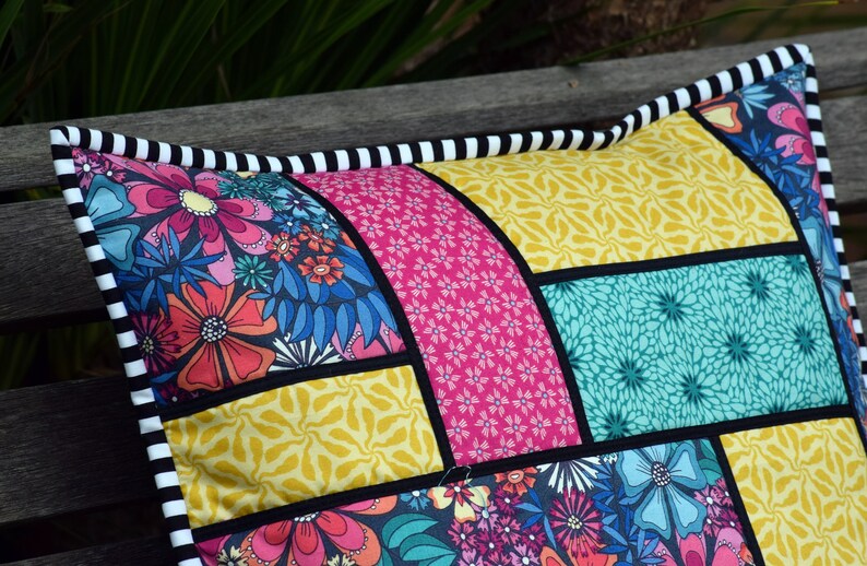 May include: A colorful patchwork pillow with a black and white striped border. The pillow is made of different fabrics with floral and geometric patterns in shades of pink, yellow, blue, and green.