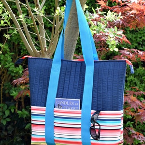 Może przedstawiać: Niebieska pikowana torba typu tote z kieszenią z materiału w paski i turkusowymi paskami. W kieszeni znajduje się książka zatytułowana "Candles on Bay Street". Okulary przeciwsłoneczne są schowane w kieszeni.