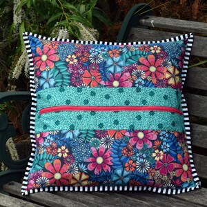 May include: A colorful floral pillow with a zippered pocket on the front. The pillow has a black and white striped border and is sitting on a wooden bench.