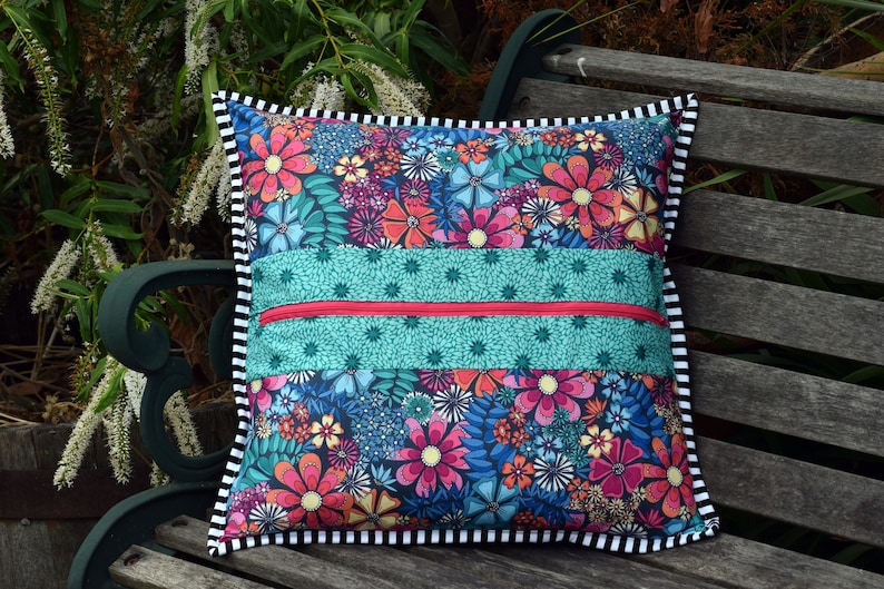 May include: A colorful floral pillow with a zippered pocket on the front. The pillow has a black and white striped border and is sitting on a wooden bench.