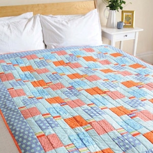 Puede incluir: Una colcha de retazos con un esquema de colores azul, naranja y rosa. La colcha está hecha de cuadrados y rectángulos de diferentes telas, y tiene un diseño simple y moderno.