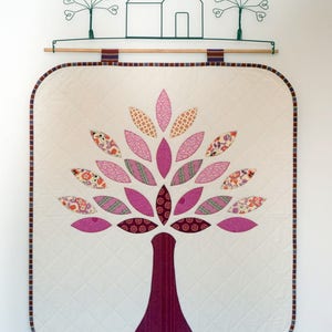 May include: A quilted wall hanging featuring a pink and purple tree with a brown trunk. The tree is surrounded by a white background with a decorative border.