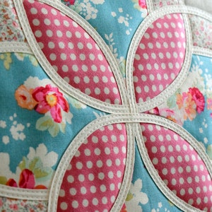 K&ouml;nnte beinhalten: Nahaufnahme eines handgefertigten Kissens mit Patchwork-Muster. Das Kissen besteht aus verschiedenen Stoffen, darunter ein blauer Blumendruck, ein rosa Tupfenmuster und ein wei&szlig;er Stoff mit Blumenmuster. Das Kissen ist mit wei&szlig;em Paspelband eingefasst.