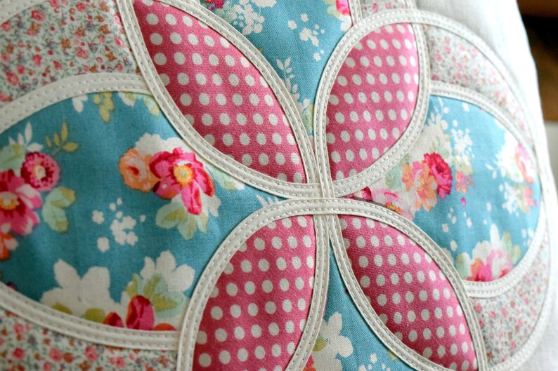 K&ouml;nnte beinhalten: Nahaufnahme eines handgefertigten Kissens mit Patchwork-Muster. Das Kissen besteht aus verschiedenen Stoffen, darunter ein blauer Blumendruck, ein rosa Tupfenmuster und ein wei&szlig;er Stoff mit Blumenmuster. Das Kissen ist mit wei&szlig;em Paspelband eingefasst.