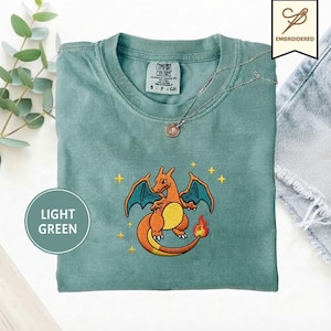 May include: A sage green crewneck t-shirt featuring an embroidered orange dragon character with blue wings and a flaming tail, surrounded by small yellow stars. The shirt has a Comfort Colors label at the neck and a silver chain necklace resting on it.