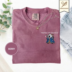 Puede incluir: Una camiseta de manga corta color baya con un bordado de una cabra vestida de mago sosteniendo una poción rosa. El texto sobre la cabra dice PREPARE FOR BEEYATLE. La camiseta tiene un cuello redondo y una etiqueta de la marca Comfort Colors.
