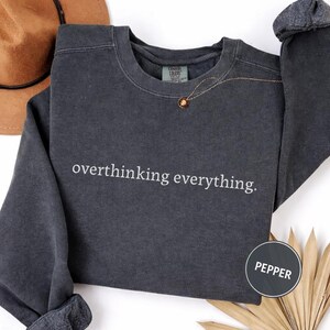 May include: A dark gray sweatshirt with the phrase "overthinking everything" in white lettering. The long-sleeved shirt has a crew neck and rolled-up sleeves. A gold necklace with a pendant is visible. A circular label reads "Pepper."