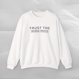 May include: A white crew neck sweatshirt with long sleeves. The text "TRUST THE CREATIVE PROCESS" is printed in black across the chest. The sweatshirt is made of a soft, comfortable material.