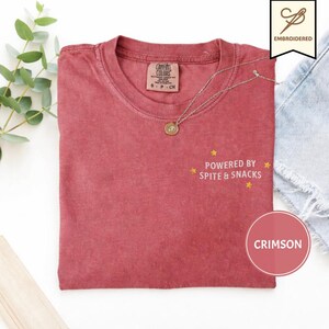 以下が含まれることがあります： クルーネックのクリムゾンカラーの半袖Tシャツ。胸元には黄色の刺繍の星と「POWERED BY SPITE & SNACKS」という文字が施されています。Tシャツの内側の襟元にはComfort Colorsのブランドラベルが付いています。
