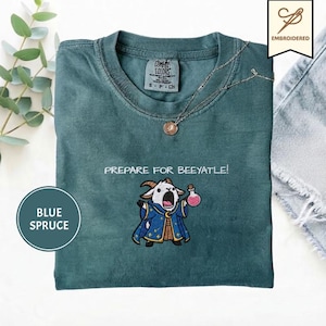 May include: A blue spruce t-shirt featuring an embroidered graphic of a goat wearing a blue wizard robe and holding a pink potion bottle. The text "PREPARE FOR BEEYATLE!" is embroidered above the goat. The shirt has a crew neckline and a tag.