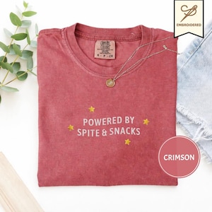 以下が含まれることがあります： クルーネックのクリムゾンカラーの半袖Tシャツ。シャツには白い刺繍で「POWERED BY SPITE & SNACKS」という文字が施され、その周囲には小さな黄色い刺繍の星が散りばめられています。衣類にはComfort Colorsのブランドラベルが付いています。
