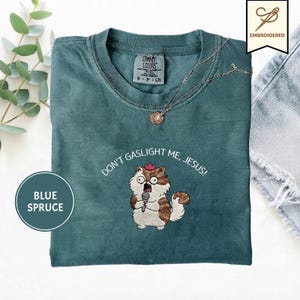 May include: A blue spruce t-shirt featuring an embroidered graphic of a fluffy brown and white cat wearing a small pink crown and holding a microphone. The text above the cat reads, DON'T GASLIGHT ME, JESUS! The shirt has a crew neckline.