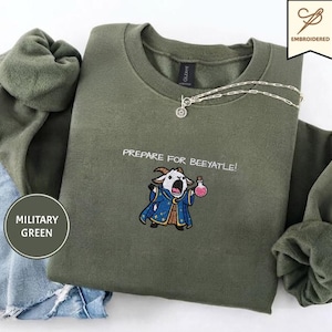 Puede incluir: Una sudadera de cuello redondo color verde militar con un bordado de una cabra vestida de mago sosteniendo una poción rosa. El texto sobre la cabra dice PREPARE FOR BEEYATLE! Un collar de cadena plateada descansa sobre el cuello de la prenda.