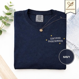 May include: A navy blue t-shirt featuring the embroidered text "CERTIFIED OVERTHINKER" in white, accented by four small yellow stars. The shirt has a crew neckline and a Comfort Colors brand label on the inside collar. A thin gold necklace rests on the fabric.